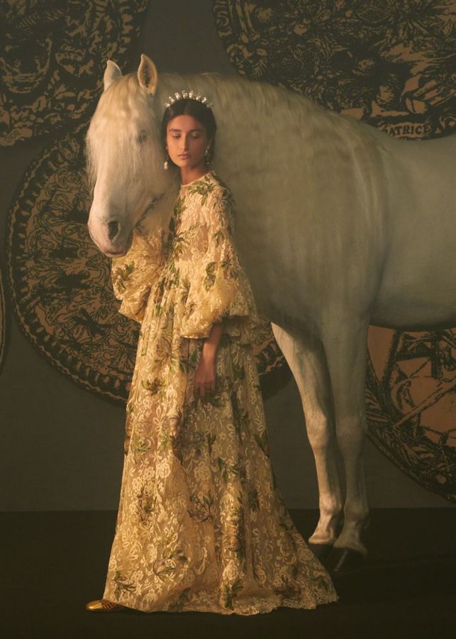 Uma modelo com vestido luxuoso de renda está de pé ao lado de um cavalo branco